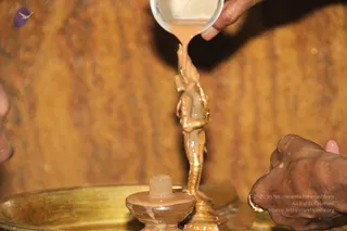 Close photo of a ritual abhishekam: a hand pours a milky liquid from a metal spoon or ladle over a small golden standing figure. A second hand with a gold ring is visible at the lower right, and a partial face appears at the far left edge. The background is a warm wooden wall.