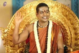 A photograph of a man seated on an ornate gold throne with carved floral scrollwork, smiling and raising his right hand in a greeting or blessing gesture. He wears a saffron-orange and gold-bordered cloth draped over his shoulder, a white flower garland, a dark rudraksha mala, a silver-and-gold ceremonial necklace, and small gold earrings. A red kumkum dot and white vibhuti marks adorn his forehead, and a small microphone clips near his face. a person is shown here, seated in a discourse setting with a red cushion behind him and a patterned fabric draped over the throne's side. Behind him a blurred blue and gold backdrop is visible.