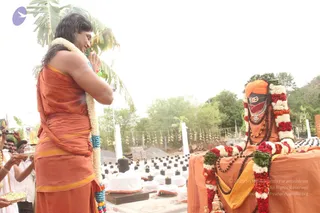 Outdoor temple-style scene with a woman in a saffron-orange sari-style wrap and a white-and-blue flower garland, hands pressed together in namaskar with eyes closed, standing in front of a seated figure draped in orange cloth, a red-and-white flower garland, a rudraksha mala and a large ornate gold necklace, wearing a saffron turban. A row of small black shivalingams sit on white cloth-covered platforms, with a banana tree, palm fronds and a lamp-filled rack behind. Another person holding a brass plate of offerings stands at the left edge.