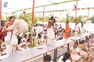 A large outdoor tent event with orange and multicoloured bunting overhead. Several young men in white dhotis with gold-bordered shoulder cloths and rudraksha malas wave white fly-whisks or fan-like cloths over a long row of seated people. Behind them, a man in an orange saffron cloth and gold necklace raises a whisk. Rows of children and adults in white sit on the ground and at long steel tables with brass plates. A tripod-mounted camera stands at the centre. The scene appears to be a ritual satsang gathering. No figure is clearly identifiable as a person from appearance cues alone.