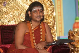 A seated man in a red-orange dhoti-style wrap and matching shawl speaks during a satsang discourse. He wears layered gold beaded necklaces, a rudraksha-style mala, gold hoop earrings, a white horizontal tilak with a red dot on the forehead, and a headset microphone. He sits on a red velvet cushioned seat in front of an ornate gold repoussé throne back, with a gold carved lion-head armrest beside him and a book or device resting on the armrest. A blue and pink backdrop is visible at right. Appearance-based cues are consistent with the page's a person reference, so this is a person identification on the throne of a discourse.