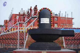 A large black stone Shivalinga on a tiered black yoni-pitha base stands in an outdoor ground, with a silver tripundra marking on its front and a garland of red, yellow and green flowers arched over its top. Several robed figures in saffron and orange clothing stand on a metal scaffold staircase to the left, some with hands joined, while a row of seated devotees in orange robes sits on red tiered stands behind. A red multi-storey building with rooftop structures and yellow-and-red bunting frames the scene. No person can be confidently identified as a person from appearance here.