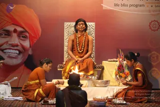 A seated figure in orange and saffron robes with a gold-yellow border sits on an ornate silver-carved throne, wearing a large rudraksha mala and gold necklace with a red tilak on the forehead. A small fire burns on a white-draped platform in front of him, with brass bowls and offering trays. Two women in orange saris with yellow borders kneel and perform rites, one holding a flower arrangement of orange and red blooms. A seated figure in dark clothing sits in the foreground with their back to the camera. A large orange-turbaned banner portrait of a smiling man and chakra graphics appear on the backdrop. Probable Swamiji: long swept-back hair, saffron robes, rudraksha mala and gold necklace on the throne, with a fire ritual in progress.