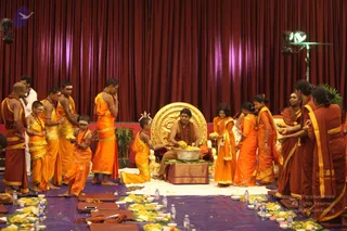 Photograph of a ritual on a stage with dark red draped curtains and a stage light rig at right. A seated man with long dark hair and a red-brown garment sits before a large gilded throne-back on a maroon cloth-draped platform, holding a metal bowl heaped with yellow flowers. Around him, young boys and women in orange and saffron garments with gold borders, some with rudraksha malas, stand and kneel, offering flowers and sweets. Floor shows banana-leaf plates with yellow flowers and sweets, water bottles, and dark wooden seats. A younger boy kneels in front, holding flowers toward the seated figure. Visible physical people number roughly 17.