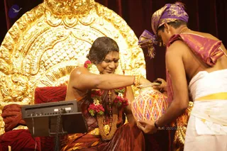 A seated figure in a maroon and brown sari with a white and red flower garland and a gold necklace receives a ritual pour from a clay pot decorated with red, yellow and white diamond patterns. A priest in a purple turban, bare-chested, with a maroon shawl and white dhoti, holds the pot. A large gold ornate throne backdrop stands behind, with a red cushioned seat and a monitor at left. Scene resembles an abhishekam pouring on a seated devotee.