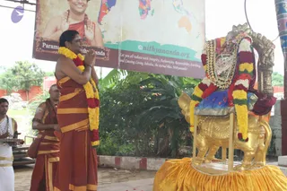 Outdoor temple-courtyard ritual photo. A man with shaved head and long hair? No — a man stands with palms pressed together in namaste, wearing a rust-orange dhoti and matching shawl draped over a bare upper body, with a fresh garland of yellow and red flowers around his neck. Behind him at right, a golden lion-form seat or vahana holds a deity murti dressed in a red and yellow flower garland, golden ornaments and a crown-like headdress. Left of centre, a priest in rust-orange cloth with white sacred marks holds a brass tray, and a man in white dhoti with rudraksha beads stands at far left. A large banner behind shows a smiling figure in saffron, a world map and the text a person and a person.org, with green and blue map colours and tropical plants in the background.