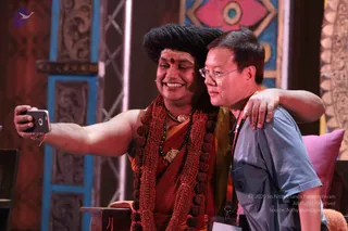 A selfie photograph on a stage with a blue and red painted backdrop. A smiling man with a long swept-back dark hairstyle, a red tilak on his forehead, a thick rudraksha mala with large beads, a red-orange cloth and a gold pendant holds a smartphone at arm's length. Beside him a man in glasses, a grey-blue short-sleeved shirt and a lanyard leans in with his arm around the first man's shoulder. Swamiji (probable) is the man in orange ochre clothing with the rudraksha garlands and long hair, shown taking a selfie with a devotee. Two people are visible.