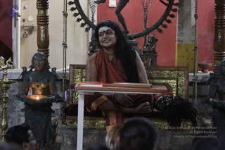 A seated figure in a red-orange robe with long dark hair and a red tilak on the forehead sits cross-legged on a gold-carved wooden seat with red cushions, smiling beside a low white table with books on it. A bronze-coloured standing lamp-bearer figure holding a lit oil lamp stands to the left, and a bronze Nataraja-style dancing figure within a flame arch appears behind. Several audience members are partially visible at the bottom edge of the frame, and red walls and a brass oil lamp frame the scene.