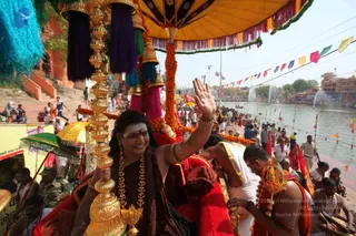 Outdoor procession at a riverside ghat with a figure seated on a red-draped palanquin under a yellow canopy with fringed trim and marigold garlands. The seated person wears a dark-maroon shawl, rudraksha-style beaded necklaces with gold ornaments, gold bangles and a tilak on the forehead, raising one hand in greeting and smiling. Golden finials and purple and pink tassels frame the palanquin. Behind, a crowd of devotees and uniformed police line the stone steps and water, where fountains spray and pennants hang on strings. A man in white and saffron-orange robes bends forward at the lower right, and a sign at the left shows Hindi text. Temple buildings sit across the water.
