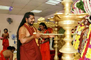 A man with long dark hair, a short beard and a red-orange dhoti and shawl stands before a large gold ceremonial oil lamp (kuthuvilakku) lit with flames, holding a tray of offerings. He wears a gold necklace, a rudraksha-style bead bracelet and armlets with a tilak on his forehead. Beside him, a younger man in orange cloth and a second robed attendant stand near a gold throne. To the right, a deity murti is adorned with flower garlands in white, yellow, pink and purple and gold jewellery. Ceiling fluorescent lights and pedestal fans are visible.