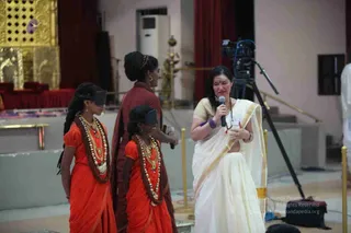 A woman in a cream and gold-bordered sari holds a microphone and speaks on a stage in a hall with gold decorative panels and a red curtain. Two young girls in orange-red dresses with white shell and beaded garlands and dark blindfolds stand at left, and an adult woman in a rust-red sari with a hair ornament walks between them holding a small object. A video camera on a tripod stands at right, and a gold railing lines the stage. Four people are visible.