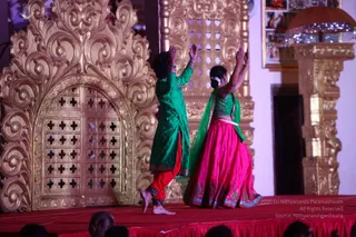 Two dancers perform on a red-draped stage in front of an ornate gold-carved wooden screen with lattice panels and a carved pillar at right. The dancer on the left wears a green kurta over red trousers with ankle bells and bare feet; the dancer on the right wears a bright pink lehenga with a green blouse and green dupatta with a flower in the hair. Both raise their arms overhead with palms open. Audience heads are visible in the foreground at the bottom of the frame, and framed pictures hang on the wall at upper right.