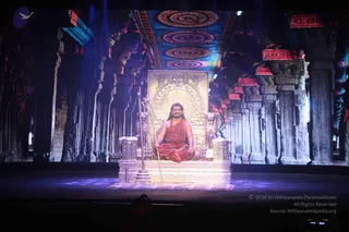 A seated man in saffron-orange robes and a red-orange shawl sits cross-legged on an ornate gold throne (peetha) on a stage, with long dark hair and a rudraksha mala around his neck. A golden staff or trident-like rod stands beside the throne, with a golden lion figure at its side. Behind him, a large stone-pillared temple hall is projected onto a screen, with colourful circular patterned ceiling panels, red hanging ornaments and blue stage lighting. In the foreground, the bottom of the frame shows the edge of the stage and a few dark silhouettes of audience members or staff. Swamiji (probable): long hair, saffron robes, rudraksha mala, seated on a throne, in the role of the presiding teacher during a stage presentation.