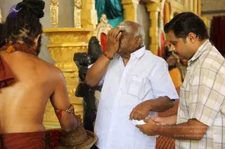 A temple interior with gilded pillars and a golden altar behind. A bare-chested priest with a long braided topknot, rudraksha beads and an orange-red cloth around his waist stands at left, holding a brass bowl on a tray with red flowers. An elderly man in a white short-sleeved shirt presses his hand to his forehead while receiving vibhuti or sacred ash, and a man in a checked shirt at right holds a small white cloth. A dark deity murti stands in the background. Context suggests a puja or darshan; the priest's cues do not clearly identify him.