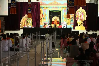 Wide photo of a large hall with a raised stage framed by maroon velvet drapes and a painted backdrop of gilded pillars and deity figures. A woman in an orange sari sits on a red-cushioned throne at centre, with a white-clad figure seated to her right and a seated person in red to her left near a cameraman. A gilded deity murti with garlands stands at left and another gilded figure with a bull-shaped base at right. Rows of seated devotees in white and orange fill the floor, with a row of steel stanchions guiding a central aisle. A small screen is visible at the upper left.