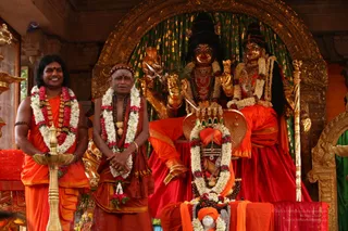 Two men stand before a golden shrine with two large deity murtis in red and gold garments, one holding a trident and a lotus-like object. The man on the left has long swept-back dark hair, orange clothing and multiple garlands, and holds a golden oil lamp. Beside him stands an older shaved-headed priest in orange, wearing a white and red flower garland, a rudraksha mala and a gold pendant, with his hands folded. A decorated lingam with flower garlands sits in front, with a shrouded orange object below. The setting is an ornate golden arch with green drapery. Partially visible text in the corner credits the photo to a person.