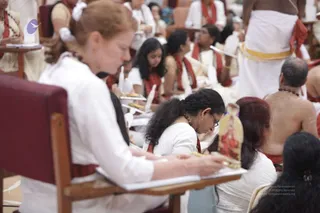 Photograph of a seated group of devotees in a hall during a ritual session. In the foreground, a woman with auburn hair in a white top sits on a maroon-backed chair, looking down and holding a pen over an open notebook. Beside her, a woman with dark curly hair and glasses wears white with a red shawl. Behind them, several people wear white garments with red shawls, and a bare-chested man with a rudraksha mala sits at right. A standing figure in white dhoti with a yellow-and-red border is visible at top right. A small gold-framed deity image sits on the tabletop at right, and small oil lamps are visible on the tables. Several people in the background hold open notebooks.