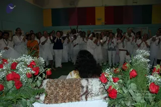 A photograph of a meditation or satsang hall with a long-haired figure seated with their back to the camera on a white-draped platform, covered by a brown and cream leopard-print fabric. Red roses and white baby's breath flowers frame the platform in the foreground. Behind, a crowd of roughly thirty people in white clothing, many wearing rudraksha or beaded necklaces, stand and applaud with raised hands. A woman in an orange sari and a woman in a navy top stand near the center. The wall behind has vertical bands of yellow, red, green and blue.