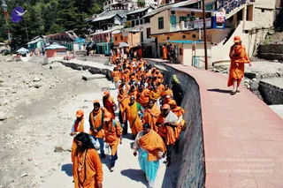 A large group of devotees in saffron-orange robes walks along a paved riverside path in a hill town, with a rocky riverbed of pale stones to the left and multistorey buildings with signboards behind. The front figure, a woman with long dark hair, wears an orange shawl, a beaded mala and a gold necklace. Others follow in orange shawls and orange caps, some carrying bags. A lone figure in orange robes walks along the red-tiled walkway on the right. The scene suggests a yatra procession through Gangotri. A sign on a building reads Bhagirathi Sadan.