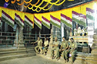 Five uniformed temple security or staff men in khaki-olive uniforms with yellow badges sit on a stone platform inside a temple hall. Festoons of yellow, white, magenta and green flower garlands hang along the ceiling beam. On the right, a shrine with floral garlands and a painted altar is visible behind a grille. Metal railings line the left side of the hall.