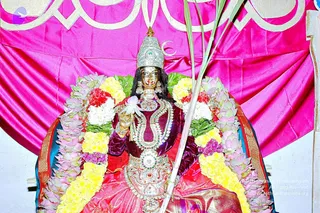 A seated deity murti, a goddess figure, adorned for Navaratri worship in a shrine set against a magenta satin backdrop with gold scalloped trim. The murti wears a silver crown with a crescent ornament, gold earrings, layered jeweled necklaces, and a deep maroon velvet blouse with a red and gold silk sari. Garlands of yellow and white flowers, pink lotus blossoms and green leaves drape around it, and a palm frond stands behind one shoulder. One hand holds a white lotus flower. No physical people are visible.