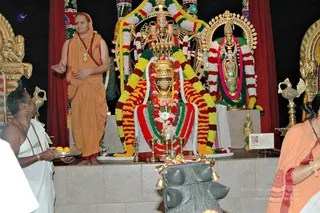 Photograph inside a Hindu temple shrine decorated with garlands of marigolds and green, red and yellow flowers. At centre sits a deity murti with a golden crown, draped in red, green and white garlands before a brass kalasha-style lamp and offerings. To the right stands a second murti in a silver-and-green garland. A man in a saffron-orange dhoti and shawl with a long gold-beaded necklace stands at left, gesturing with open hands. Beside him a man in white ritual cloth holds a silver tray of fruit and an oil lamp. A woman in an orange sari with a pink border stands at the right edge. Gold lotus-style shrine ornaments frame the altar.