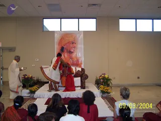 A photograph of a ceremony on a raised white-draped stage in a bright hall with beige walls and tall windows. A man in white dhoti and shawl, with grey hair, stands at the left holding a silver plate. On the stage, a figure in white dhoti and red shawl kneels or bends beside a seated figure in saffron-orange robes wearing a garland of flowers and a red cloth. Behind them hangs a large orange-toned portrait of a man wearing an orange turban. Flower arrangements in yellow and purple sit at the stage corners, and a seated audience in red, black and white clothing faces the stage from the lower foreground.