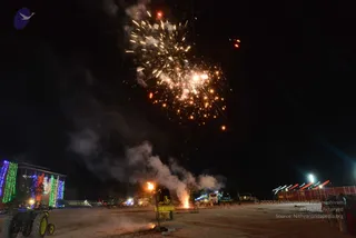 Night photograph of an outdoor ground at an ashram with a fireworks display bursting into bright sparks in the dark sky above. Smoke drifts low across the open dirt field. A small fire glows near a yellow machine at centre, and a tractor sits at far left beside a multi-storey building wrapped in green, blue and red lights. Vehicles and fence lights line the right edge. No people are clearly visible; a few small figures appear distant near the right fence.