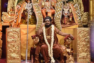A man seated on an ornate golden throne with lion-head armrests, wearing a rust-brown saffron-toned robe and shawl, a white jasmine garland, a rudraksha-style gold pendant necklace and multiple gold chains, and a bangle on his arm. He has long dark hair, a beard and white ash markings on his forehead, and rests one hand near his face. Behind him are gilded deity murti figures with crowns and jewelled ornaments, and a silver trident stands to his left against a gold-embroidered backdrop. The setting is a richly decorated shrine-like hall. Scale: one person is physically present in the scene.