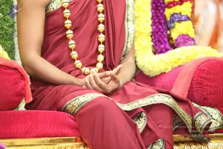 Close photo of a seated figure on a red cushioned throne with gold-edged seat, wearing a maroon-red draped garment with gold brocade borders. A long necklace of large gold beads hangs across the chest. The hands rest in a meditation mudra on the lap. Behind the figure are garlands of yellow and purple marigold-type flowers and green foliage. The lower body and face are cropped out of frame. The figure appears to be a person (in_person) by the saffron-red robe, ceremonial gold necklace and throne setting.