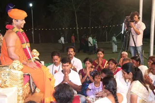 Night outdoor satsang with a man seated on a gold lion-armrested throne, wearing an orange-saffron dhoti-style garment, an orange turban and a long red and white flower garland; he smiles while facing a seated group. Many devotees sit on a green mat with hands folded in anjali, including men in white shirts, a woman in a maroon sari, a woman in a red-bordered sari, a girl in a pink top, and women in white saris. A camera operator in a pink shirt with headphones films from a tripod at right. A string of lights and trees appear behind, with a few people standing in the background. Several people are visible, and a copper-coloured vessel sits near the front, with a white candle or lamp on a stool beside the throne.