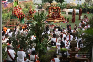 Outdoor garden satsang in a courtyard with lawn, tropical plants and a stone fountain in the foreground. Seated on a golden throne decorated with red and orange marigold garlands is a figure in red-orange robes with a red shawl, flanked by golden parasols. Two further garlanded figures sit on a raised green-draped platform at left, and a monk in orange robes stands at right. Dozens of devotees in white clothes with red-orange shawls sit on the grass facing the dais, with some seated on chairs at the back. Brass lamps and offerings are arranged on a mat before the platform.