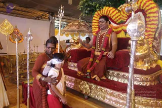 A seated man in an orange-red dhoti and shawl, wearing rudraksha and ceremonial beaded necklaces, a red and orange flower garland, and gold bangles, raises his hand on a gilded throne with red cushions and a golden lion-shaped armrest. In front, a devotee in orange-red robes with a sandalwood-coloured shawl guides a blindfolded young boy in white, bare-footed, toward the throne. Silver trident staffs, a gold-and-red ornate parasol, brass lamps and a golden decorative banner stand in the hall, which has a white draped backdrop and a fabric ceiling. Appearance-based a person seated on the throne performing a samaya deeksha initiation ritual, with a blindfolded initiate being brought forward.