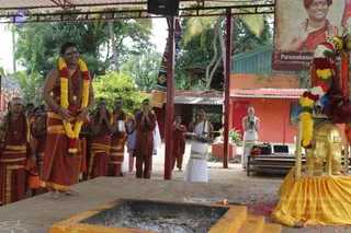Outdoor temple ceremony under a corrugated roof at a dhwajarohanam flag-hoisting event. In the foreground, a bare-chested man in saffron-orange dhoti with yellow borders stands with palms joined, wearing a large marigold and red flower garland, a rudraksha mala, and a sacred-thread-style necklace; his pose suggests a presiding figure. Behind him, several men in orange and saffron robes stand with hands folded, some holding small brass vessels. A woman in a white sari with gold border holds a silver tray. At right, a golden elephant-shaped base holds a red and gold cloth and flower garlands. A square fire pit with offerings sits in the foreground, and a banner shows a portrait labelled "Paramahamsa Nithyananda" in saffron robes. Scattered flower petals cover the floor.