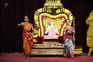 Two young female dancers perform on a red stage before a heart-shaped throne decorated with yellow, red and white flower garlands and a gold canopy. The dancer on the left wears a mustard-orange and red saree with gold jewellery and bangles and gestures with her hands. The dancer on the right wears a grey and red saree with gold necklace and bangles and raises her hands in a mudra. Behind them a printed portrait of a seated figure in saffron-red robes and rudraksha malas appears on the throne backdrop, flanked by a golden lion-form sculpture and a tiger-skin cushion. Maroon curtains frame the stage.