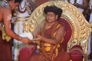 A seated figure on an ornate gold throne with red cushions receives a ritual offering from a bare-chested attendant in a rust-coloured dhoti with white ash markings on his arms. The seated man wears a rust and yellow shawl, a long rudraksha mala and a gold chain, with a red bindi on his forehead and eyes closed. Two white-clad priests stand behind, one holding a small plate. The attendant holds a white cloth-wrapped coconut over a steel bowl. Floral garlands and a microphone are visible. a person depicted in the role of the seated honoured guest during a temple ceremony.