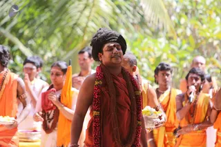 Outdoor procession under palm and leafy trees. A central man in a rust-red robe wears a long red and yellow flower garland, a rudraksha-style brown bead garland, and a dark swept-up head covering with a red-and-white tilak on his forehead. Around him, devotees in saffron-orange dhotis and shawls carry silver trays of white and yellow flowers, and one holds a microphone. Several people walk with hands in anjali, and a saffron umbrella is visible at left.