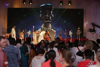 A photograph of a large stage event with a dark star-field backdrop and a large stone Shiva-type murti with a cobra hood above a pedestal. On the red-draped stage, a man in a white dhoti and gold-trimmed shawl stands before a seated person in saffron and red garments, while another man in white kneels and a woman in white and red is nearby; trident standards flank the platform. A dense audience in the foreground, many in white and pastel clothes, watches. A film camera on a crane is at right. The visible stage figures are not confidently matched to any identity, so no candidate is asserted.