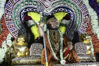 A seated man in a decorated procession setting holds a silver trident in his right hand. He wears a gold crown, a brown-orange saffron shawl over a leopard-print garment, a heavy gold and rudraksha-style necklace, and a white flower garland. Two golden lion sculptures flank a leopard-print throne, and a large heart-shaped backdrop of white, yellow and red flowers with embroidered mirror work surrounds him. The scene resembles a festival procession throne.