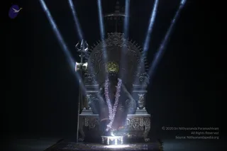 A figure sits on an ornate silver throne (peetha) with carved floral and lion-head details, lit from above by bright beams against a dark backdrop. The seated figure wears a gold crown, a pink-and-white flower garland, and dark wrapped garments. A silver trident stands to the left of the throne, and a small lit pedestal with a white glow sits at the base on a patterned rug. The scene is a dramatic staged darshan setting.