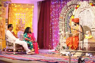 Indoor satsang on an ornate stage. On the right, a bearded man in an orange-saffron dhoti and shawl with a saffron turban and beaded necklaces sits on a silver-and-patterned throne, raising one hand as he speaks into a microphone. Two guests sit on silver chairs to the left: a man in white kurta with an orange-red shawl, and a woman in a teal-and-green embroidered kurta with a pink dupatta, holding a microphone. Behind them stands a gold-ornate carved pillar with a female deity murti, strings of purple fairy lights, a maroon curtain and a colourful woven rug. Several people appear across the frame.