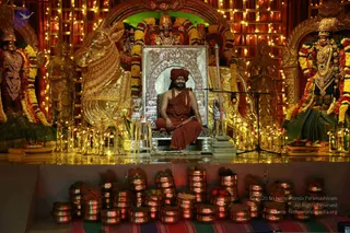 A seated man in a saffron-red turban, saffron-red robes and a long rudraksha mala sits on a silver-framed throne-like seat in front of a golden horse-shaped sculpture and tiered oil lamps. Behind him, gold statues and floral garlands frame a shrine-like stage, with a microphone stand in front. Rows of brass pots and kalasha-style vessels with offerings fill the lower platform, and strings of lights glow in the background.