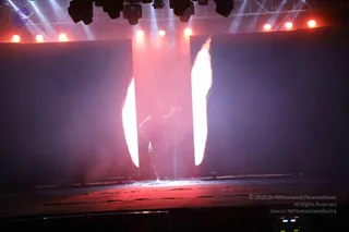 A dimly lit stage with red and white spotlights overhead, two dark tall panels flanking the centre, and bright white vertical light strips glowing beside each panel. Hazy red-tinted smoke fills the central space, and a single dark figure stands in silhouette near the middle of the stage, partly obscured by the haze, with thin vertical stage elements behind. The floor reflects the red light. The image is a stage performance photograph, not a formation of people.