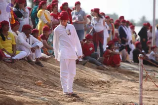 A photograph of an outdoor gathering on a dry dirt slope. In the centre a young boy in white kurta and pyjama-style trousers and a red turban walks downhill barefoot in sandals, a small white badge on his chest. Behind him many people in red headwraps sit and stand on the slope, including a woman in a yellow kurta, a man in white with a red head covering, and women in red and yellow saris. A white post with a red rope stands at right. Visible people number roughly twenty to thirty.