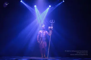 A man stands alone on a dark stage under blue and green spotlights, bare-chested with a crescent ornament on his head and a long ornate necklace and sash. He wears rust-red draped lower garments and ankle bands, and holds a tall silver trident with a wrapped shaft, styled as a Shiva-like figure. The scene is a theatrical performance, possibly a culturals segment, lit with dramatic beams against a black backdrop.