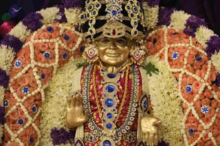 A golden statue of a figure is adorned for worship, with a tall ornate crown studded with blue stones and a red centre gem, and jewelled earrings. The statue wears a red and gold garment with a layered necklace of blue gems, pearls and a gold chain. Its right hand is raised in abhaya mudra and the left hand rests open. The figure is framed by orange and pink roses, white jasmine garlands and purple flowers forming a decorated arch, resembling a murti being honoured during a puja or darshan. No living people are visible.