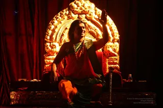 A seated man on a dark stage raises his right hand in blessing while smiling. He wears an orange-red drape over a green-and-gold bordered garment and a long rudraksha mala. Behind him, a large ornate golden arch-shaped throne glows against deep red velvet curtains. A small table with a water bottle sits at right, and the figure is lit in warm golden light. a person, seated on a throne in an orange robe with rudraksha mala, giving blessing during a satsang-style stage setting.