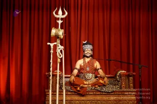 A seated man sits cross-legged on an ornate carved wooden throne draped with a leopard-print cloth, in front of deep red curtains. He wears a saffron-orange dhoti and a matching orange wrap, a leopard-print headband with a small crescent ornament, layered rudraksha malas and a pendant necklace, and beaded wristbands. A silver trident with a damaru drum hangs beside him, and a microphone stands at his right. He smiles with eyes closed in a satsang setting.