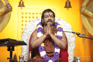 A man seated on an ornate white and silver throne with red cushioning, against a yellow backdrop with golden bell and wing motifs. He wears a rust-orange robe, a rudraksha mala with a ceremonial gold pendant, and a long garland of purple and white flowers. He has white horizontal tilak marks and a red bindi on his forehead, and his palms are pressed together in namaste while a microphone stands before him. A keyboard sits at the left edge. Swamiji (probable) is shown in this photo, seated in the central position of a satsang-style discourse, with long swept-back hair, rudraksha malas and ornate gold adornments.