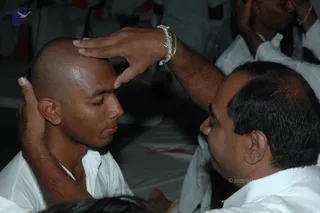 A photograph of an initiation moment in a seated gathering. A shaved-headed young man in a white shirt sits with eyes closed while an older man in white, with a silver bracelet on his raised wrist, presses his palm on the young man's forehead and supports the back of his head. Other seated men in white clothes are visible behind them, some with hands raised to their own heads. The setting is dim, with white sheets or cloths on the floor. No a person candidate is clearly identifiable by appearance, and the people shown are devotees in white.